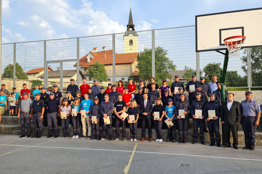 Skupinska fotografija ministra Tonina z novimi operativnimi gasilci na športnem igrišču, v ozadju hiše in cerkveni zvonik
