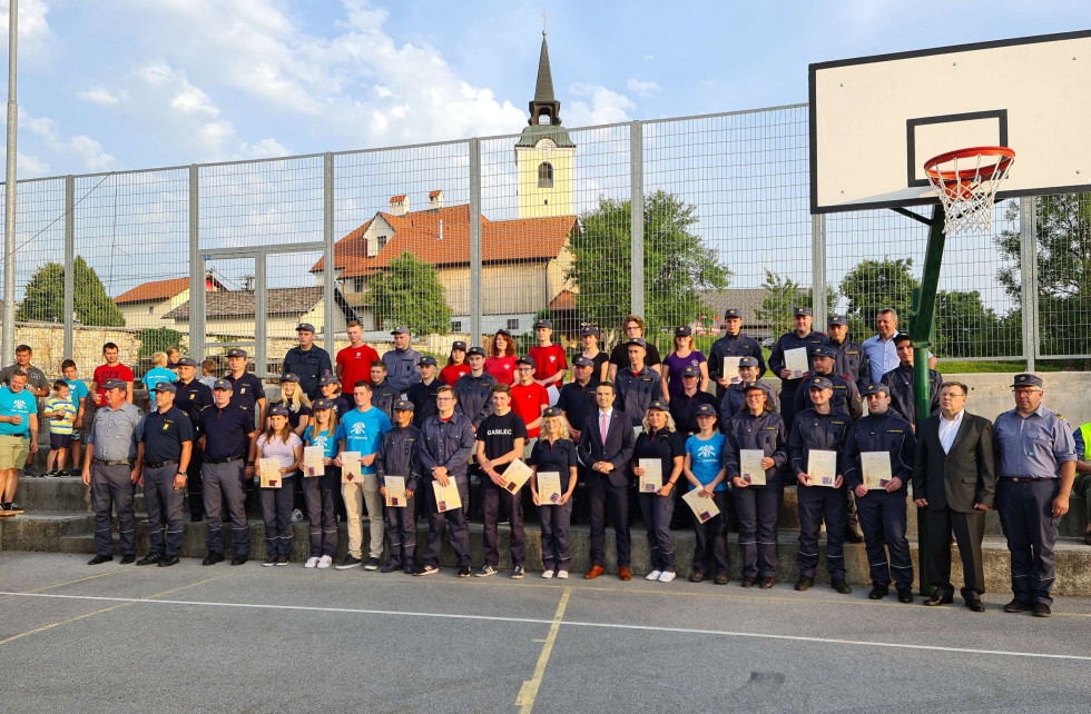 Skupinska fotografija ministra Tonina z novimi operativnimi gasilci na športnem igrišču, v ozadju hiše in cerkveni zvonik