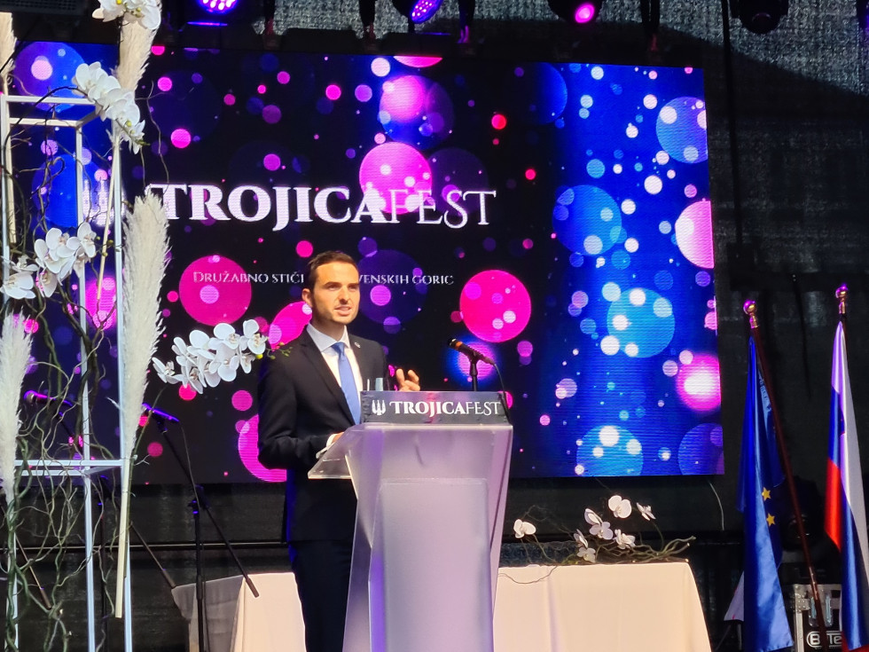 Minister za obrambo mag. Matej Tonin stoji za govorniškim pultom z napisom Trojicafest, ki je izpisano tudi v ozadju na modri podlagi z rožnatimi krogi