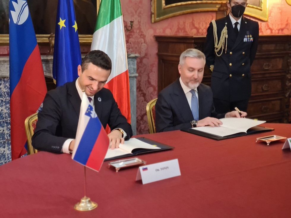 Ministra podpisujeta dogovor. V ozadju so zastave Slovenije, EU in Italije.