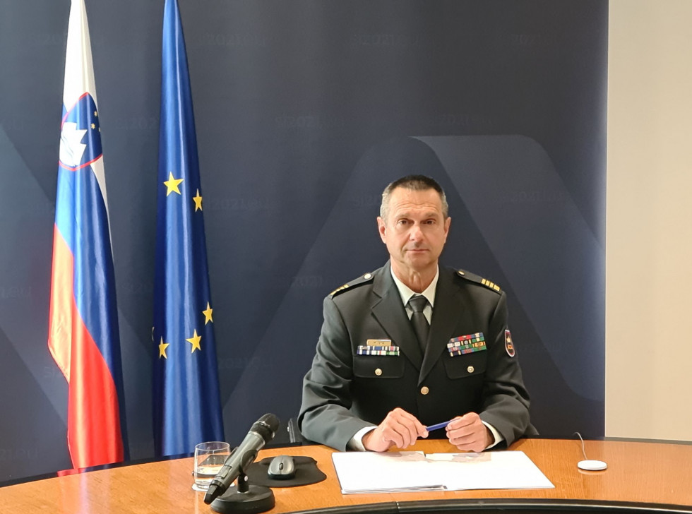 Polkovnik sedi za konferenčno mizo z gradivom pred seboj. V ozadju je temno moder pano predsedovanja in zastavi Slovenije ter EU.