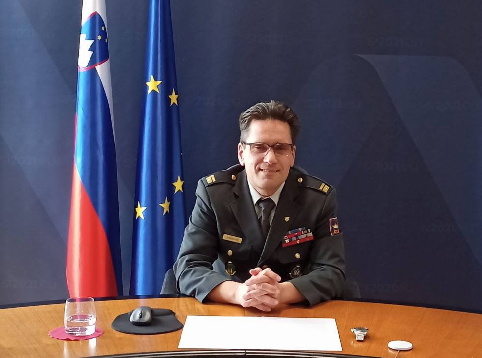 Major med novinarsko konferenco sedi za mizo z gradivom pred seboj. Za njim je temno modro ozadje in zastavi Slovenije ter EU.