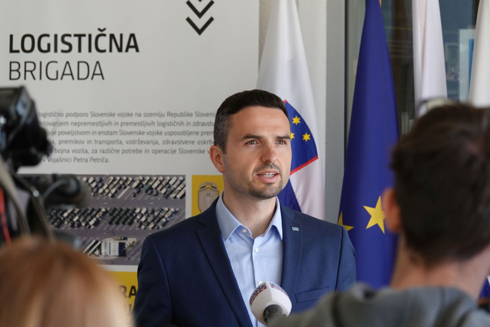 Minister pred mikrofoni na novinarski konferenci. Zadaj so zastave Slovenije, EU ter razstavni panoji 