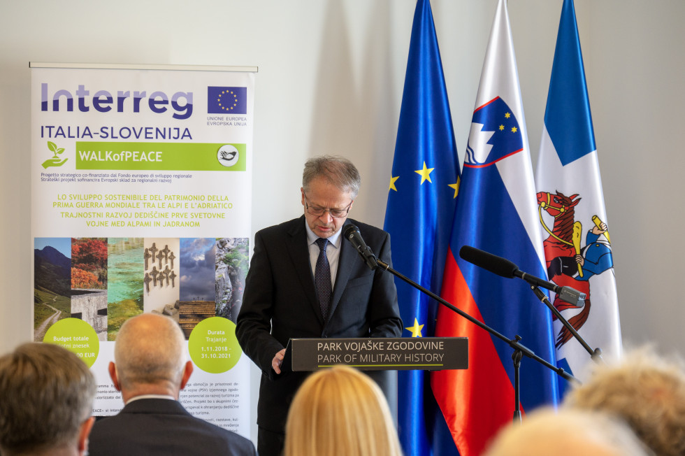 Sekretar med govorom za govornico. Za njim so zastave Slovenije, EU in občine Pivka ter pano razstave