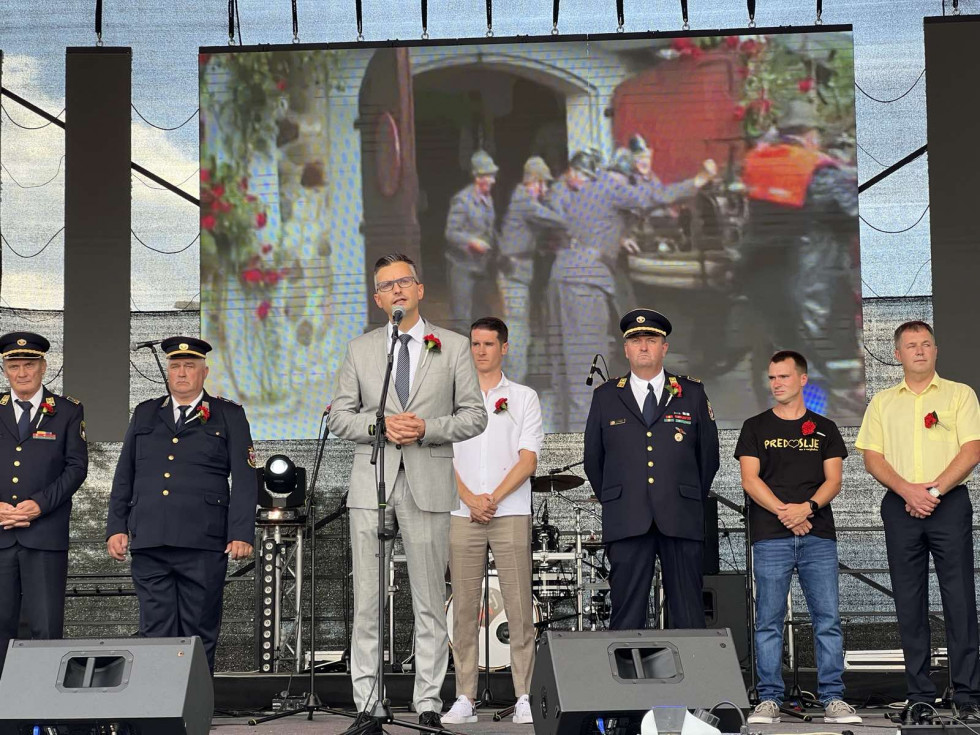 Minister med nagovorom stoji na odru. Za njim so gasilci, predstavniki društva. Na platnu je projekcija zgodovine društva