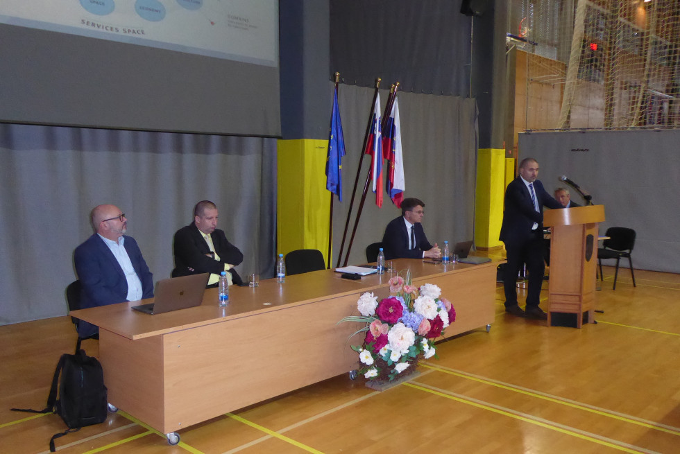 Vodja vaje med govorom stoji za govornico, poleg pa za mizo sedijo člani vodstva vaje. V ozadju  so zastave Slovenije in EU