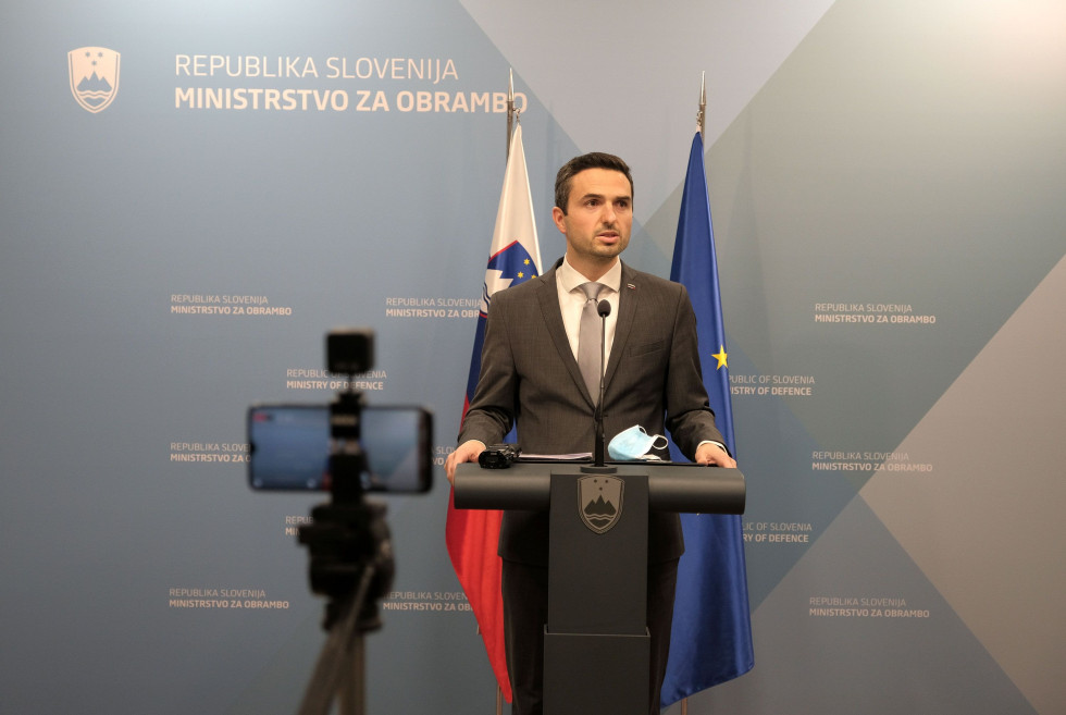 Minister za govornico med novinarsko konferenco. Za njim je moder pano ministrstva in zastavi Slovenije ter EU