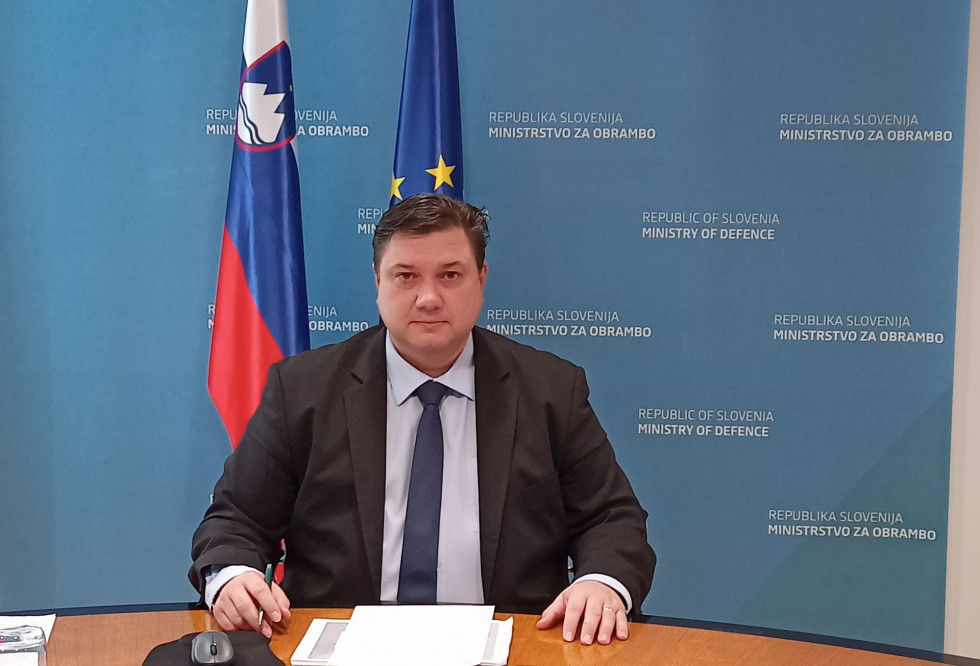 Direktor sedi za mizo med video konferenco. Pred seboj ima gradivo, v ozadju pa je moder pano ministrstva in zastavi Slovenije ter EU.