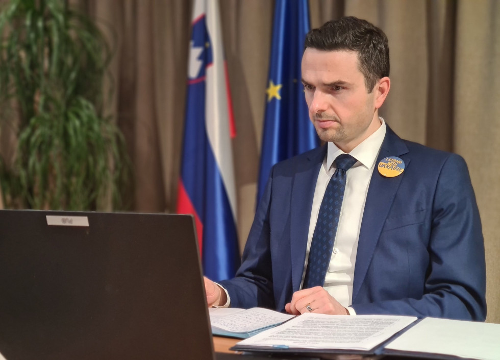 Minister sedi pred računalnikom-Zadaj sta zastavi Slovenije in EU.