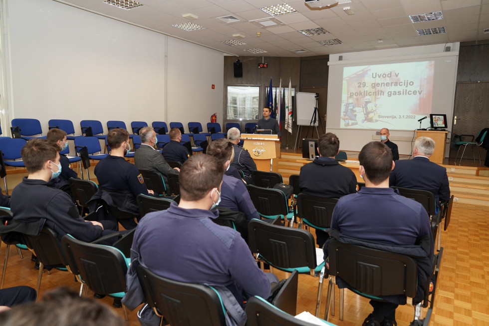 Minister stoji za govornico pred avditorijem polnim gasilcev. V ozadju je platno s projekcijo.