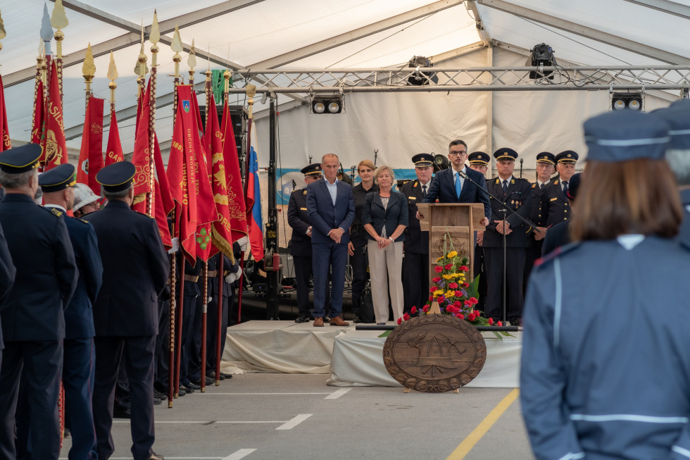 Minister med govorom za govornico. Ob njem stojijo udeleženci proslave, ob strani pa gasilci s praporji društev