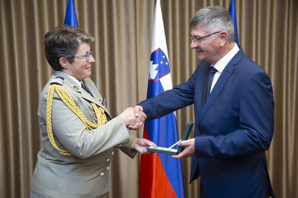 Sekretar izroča medaljo atašejki.Za njima so zastave Slovenije, Francije in EU