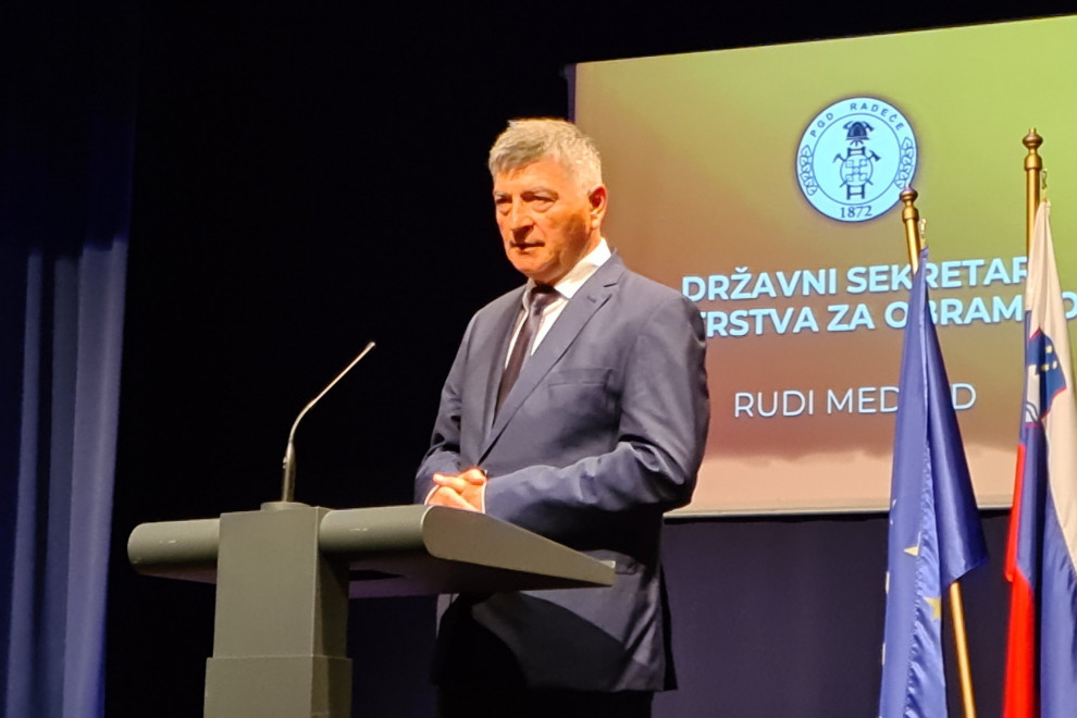 Sekretar stoji za govornico med govorom. V ozadju je projekcija z znakom društva in njegovim imenom