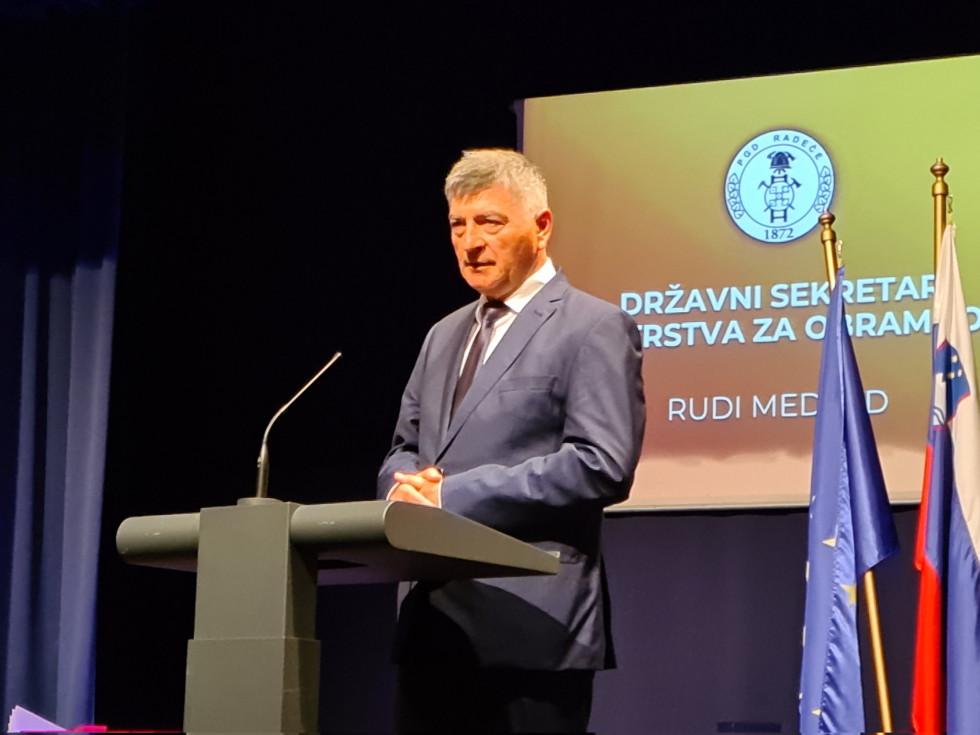 Sekretar stoji za govornico med govorom. V ozadju je projekcija z znakom društva in njegovim imenom