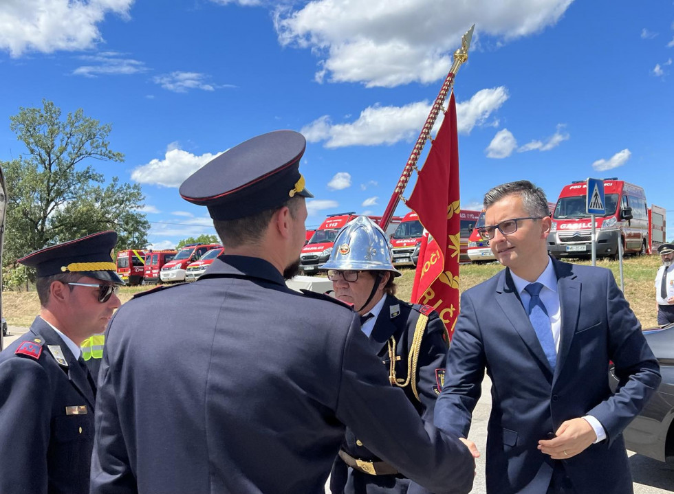 Minister se rokuje z gasilci. Za nim so gasilska vozila in gasilka s praporjem društva