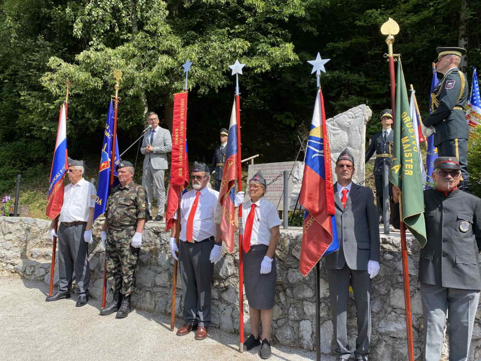 Minister med govorom stoji na odru. Pod odrom so v vrsti udeleženci proslave s praporji