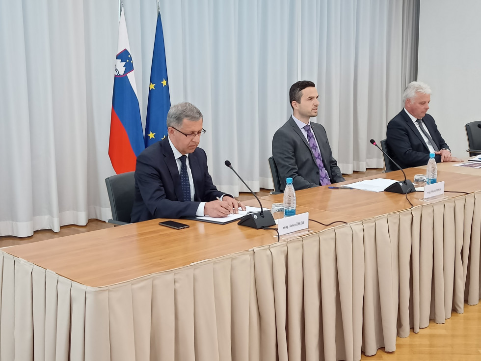 Minister, sekretar in direktor sedijo za konferenčno mizo z mikrofoni pred seboj. Za njimi sta zastavi Slovenije in EU.