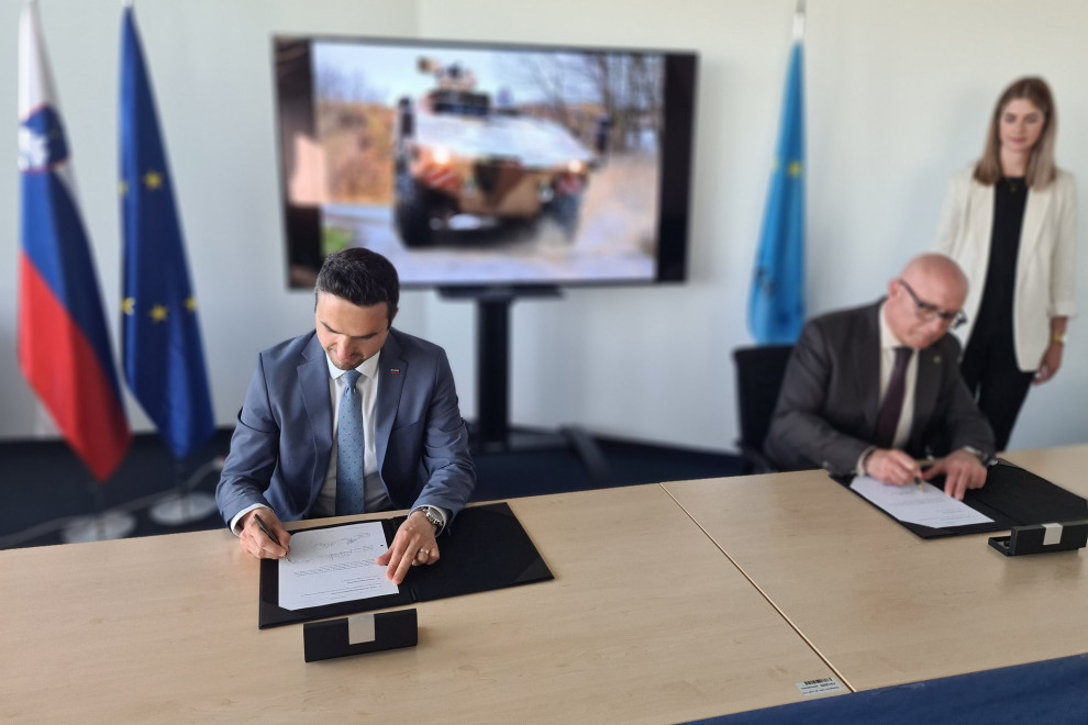 The Minister and the Director of OCCAR sign the documents seated at a table. Behind is a screen with a picture of a boxer vehicle