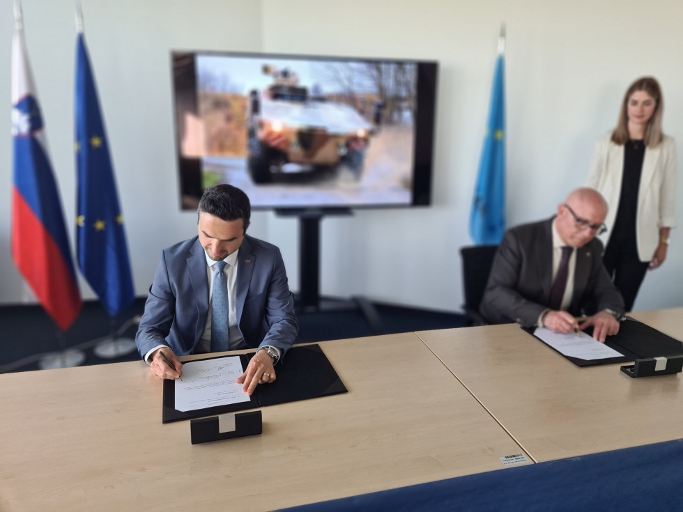 The Minister and the Director of OCCAR sign the documents seated at a table. Behind is a screen with a picture of a boxer vehicle