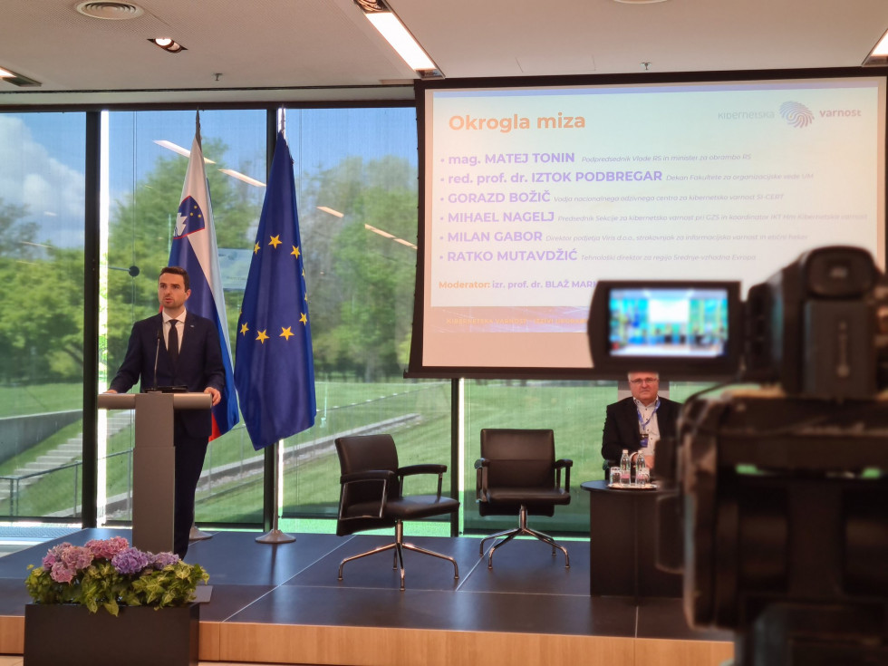 Minister stoji za govornico. Za njim sta zastavi Slovenije in EU ter platno s projekcijo,