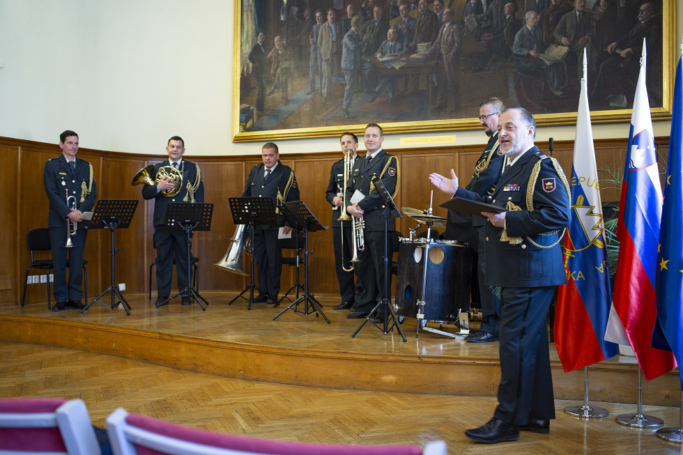 Člani orkestra in dirigent pred nastopom. Zadaj so zastave Slovenije in EU, na stani pa velika umetniška slika