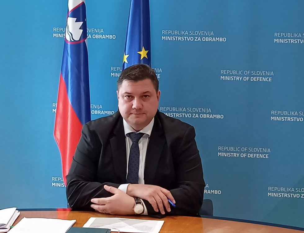 Direktor sedi za mizo z gradivom za video konferenco pred seboj. V ozadju je moder pano ministrstva in zastavi Slovenije ter EU