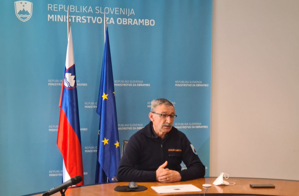 Poveljnik med videokonferenco sedi za mizo. Za njim je moder pano ministrstva in zastavi Slovenije ter EU