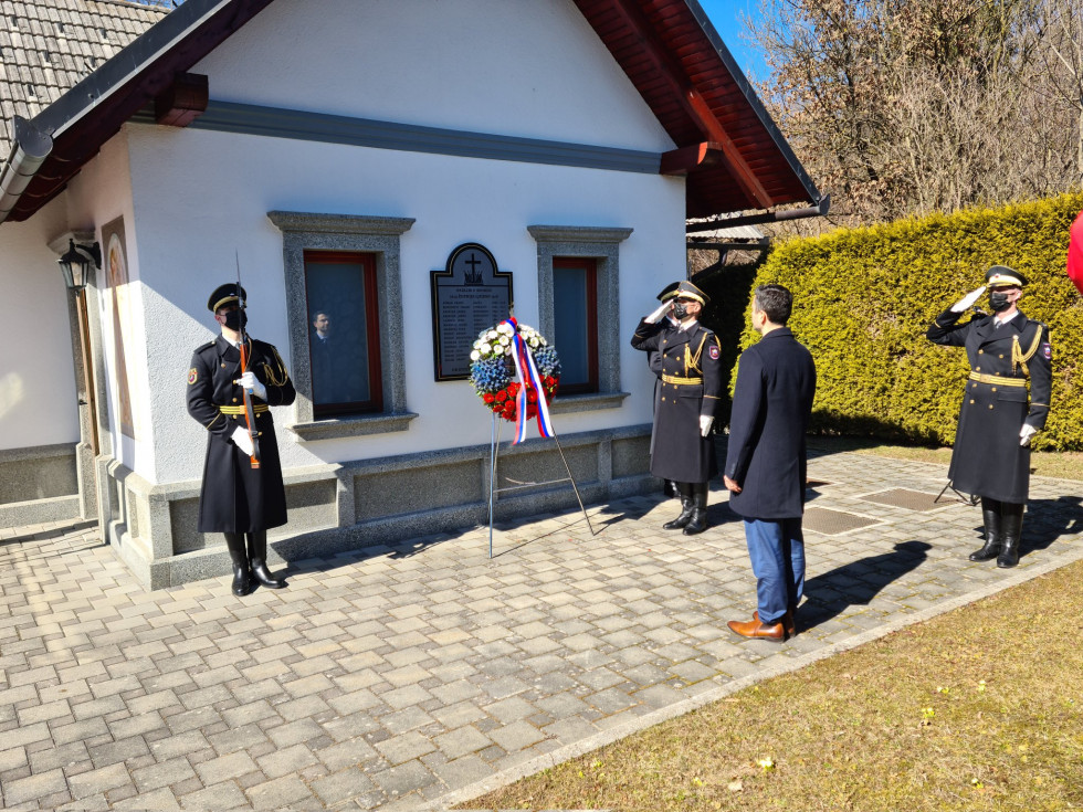 Minister stoji pred hišo s spomenikom padlim v 1. svetovni vojni in vencem, ki ga je položil. Na vsaki strani stojita gardista Slovenske vojske