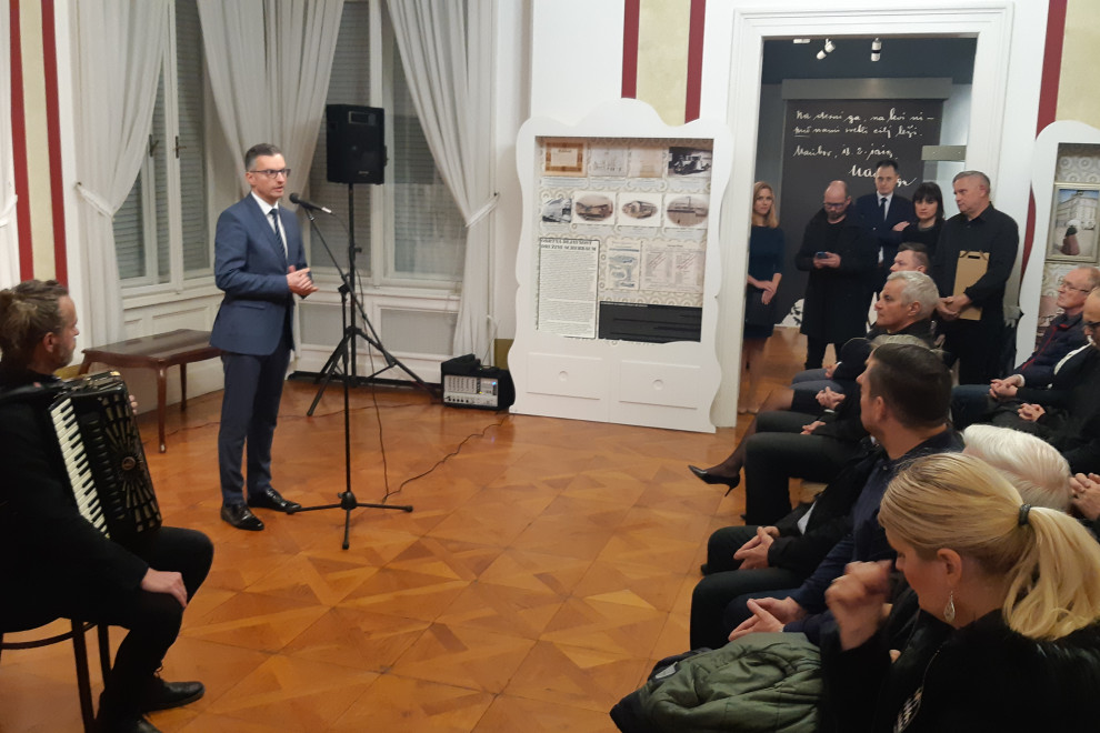 Minister stoji pred mikrofonom v muzejski dvorani pred občinstvom ob otvoritvi razstave