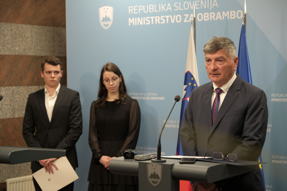 Sekretar stoji za govornico. Desno od njega stojita gosta, za njimi sta zastavi Slovenije in EU ter moder pano ministrstva