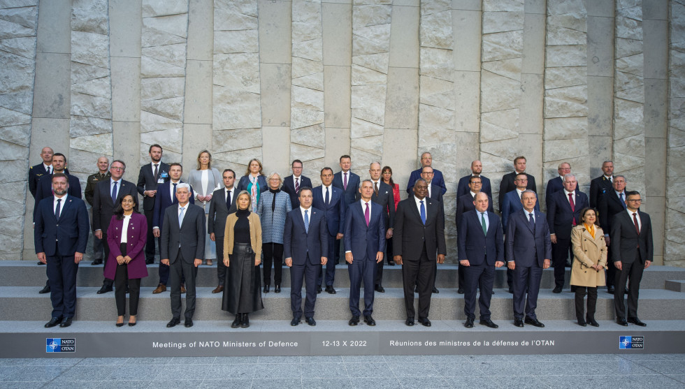 Group photo of ministers. They are standing on low steps, with NATO flags and a high stone wall behind them.