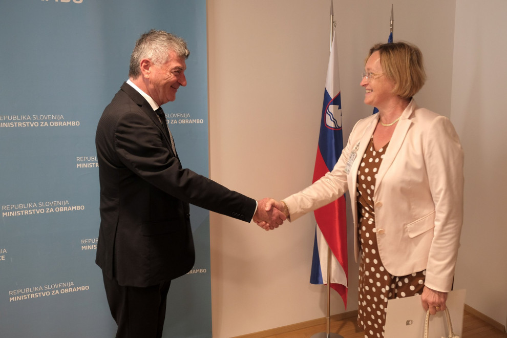 Sekretar in predstavnica se rokujeta pred zastavama Slovenije in EU