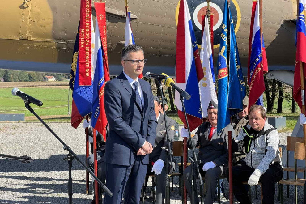 Minister med govorom stoji pred mikrofonom. V ozadju sedijo udeleženci slovesnosti in stoji letalo DC-3
