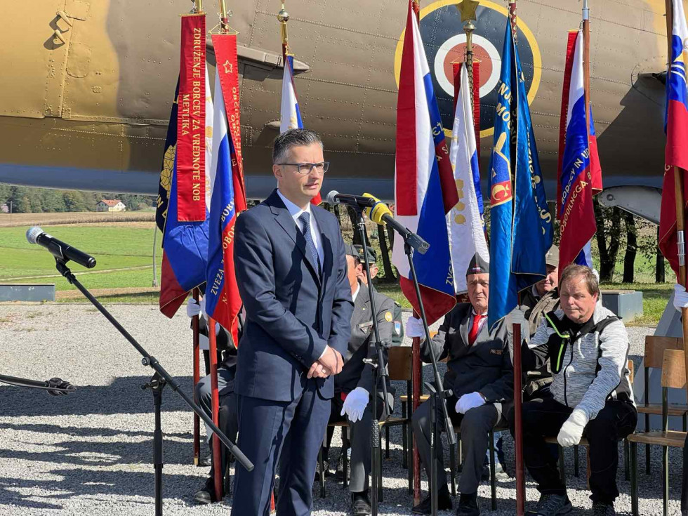 Minister med govorom stoji pred mikrofonom. V ozadju sedijo udeleženci slovesnosti in stoji letalo DC-3