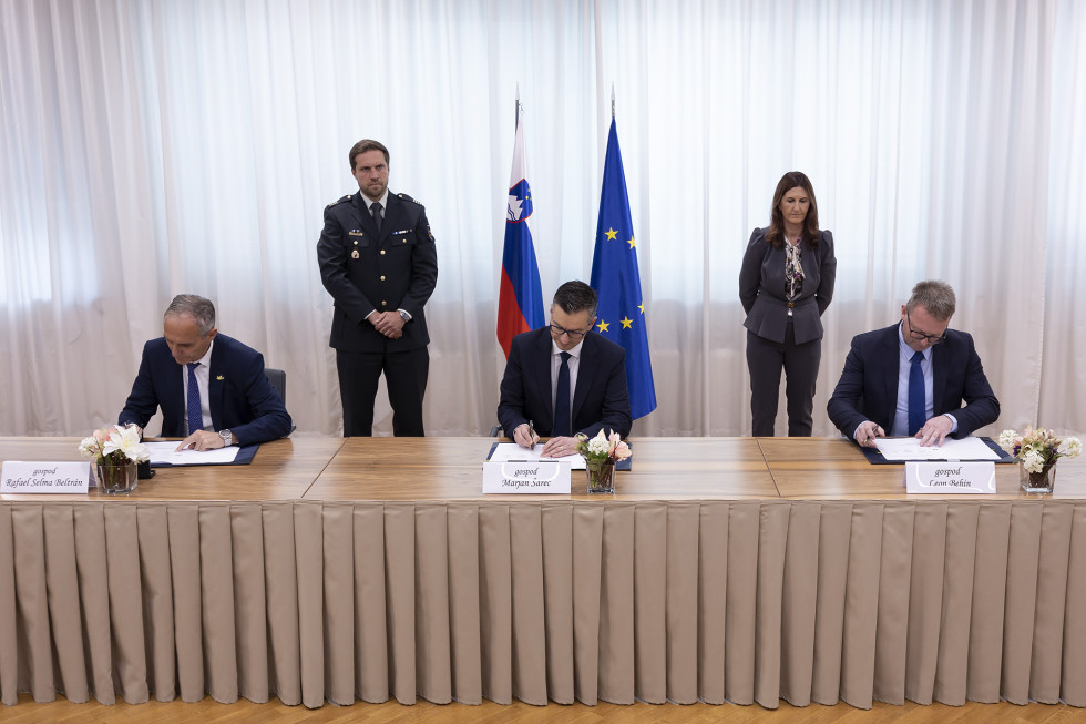 Podpisniki med podpisom sedijo za dolgo mizo. Za njimi so predstavniki protokola in zastavi Slovenije ter EU