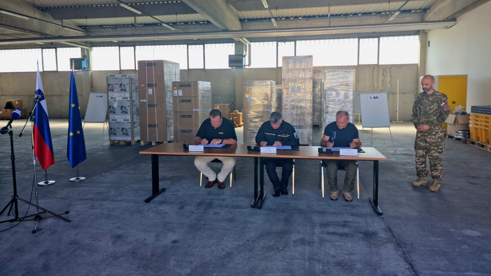 Podpisniki sedijo za mizo v skladišču in podpisujejo pogodbo, desno stoji pripadnik Slovenske vojske, v ozadju je oprema v embalaži.