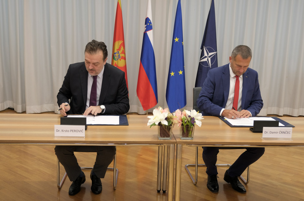 The two secretaries sit at the table and sign the agreement. Behind them are the flags of Montenegro, Slovenia, the EU and NATO.