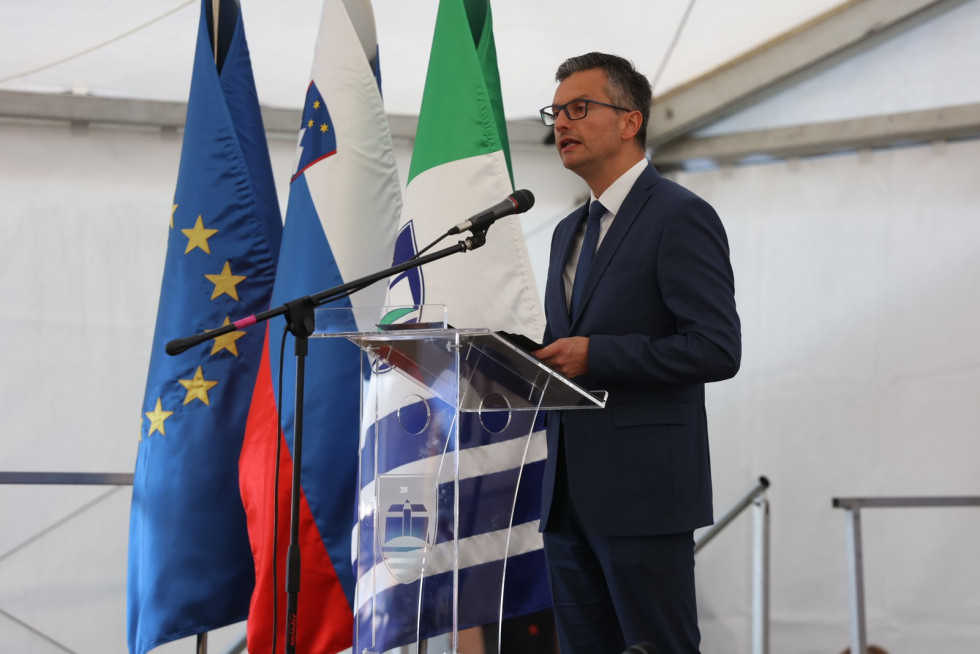 Minister stoji za govorniškim odrom in nagovarja zbrano občinstvo, levo zastave Evrope, Slovenije in Občine Lendava.