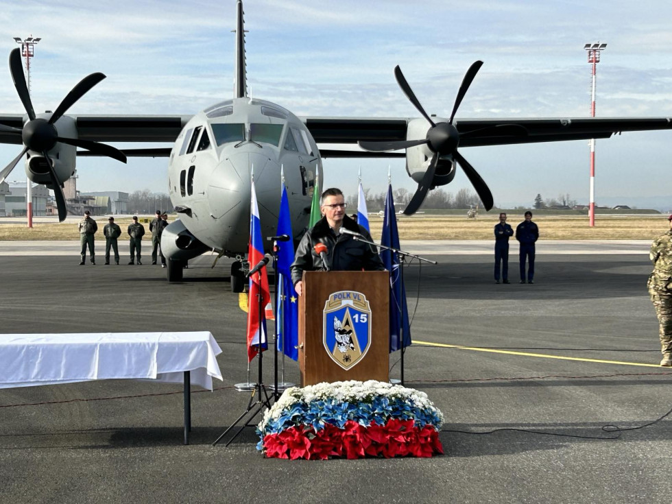 Minister med govorom za govornico na letališki stezi. Za njim je letalo Spartan