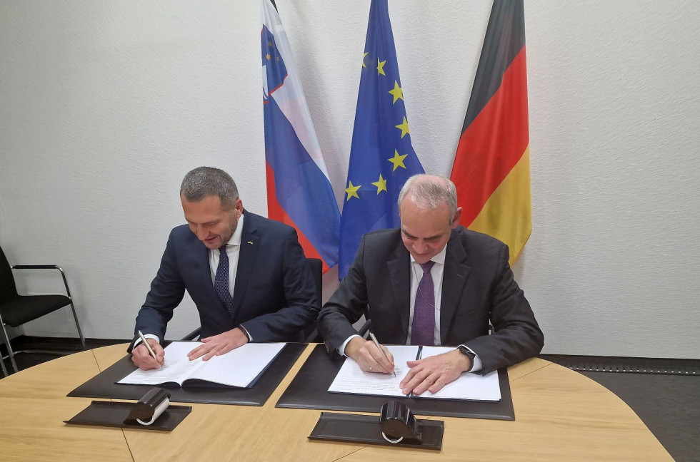 The two secretaries sit at the table while signing. Behind them are the flags of Slovenia, the EU and Germany