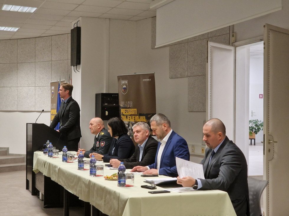 The conference participants sit at a long meeting table. The moderator stands next to them behind the podium