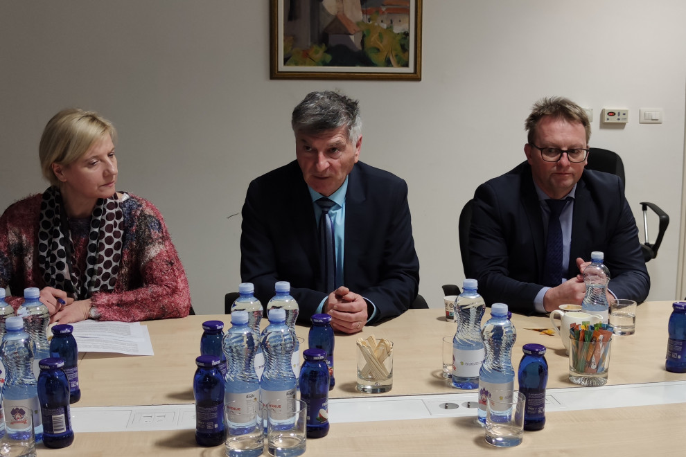 Sekretar in direktor med srečanjem sedita za sejno mizo. Za njima na zidu visi umetniška slika