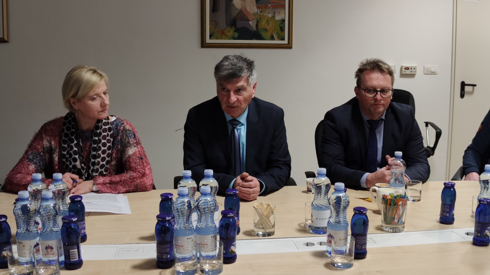 Sekretar in direktor med srečanjem sedita za sejno mizo. Za njima na zidu visi umetniška slika