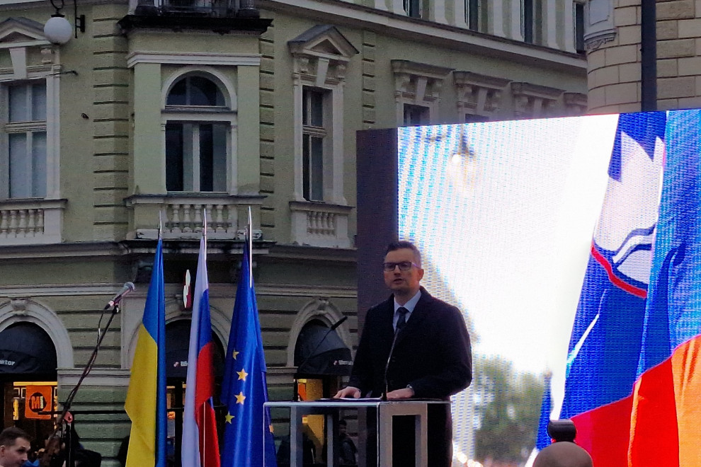 Minister med govorom stoji na odru. Za njim je velik zaslon, in zastave Slovenije, Ukraijne ter EU
