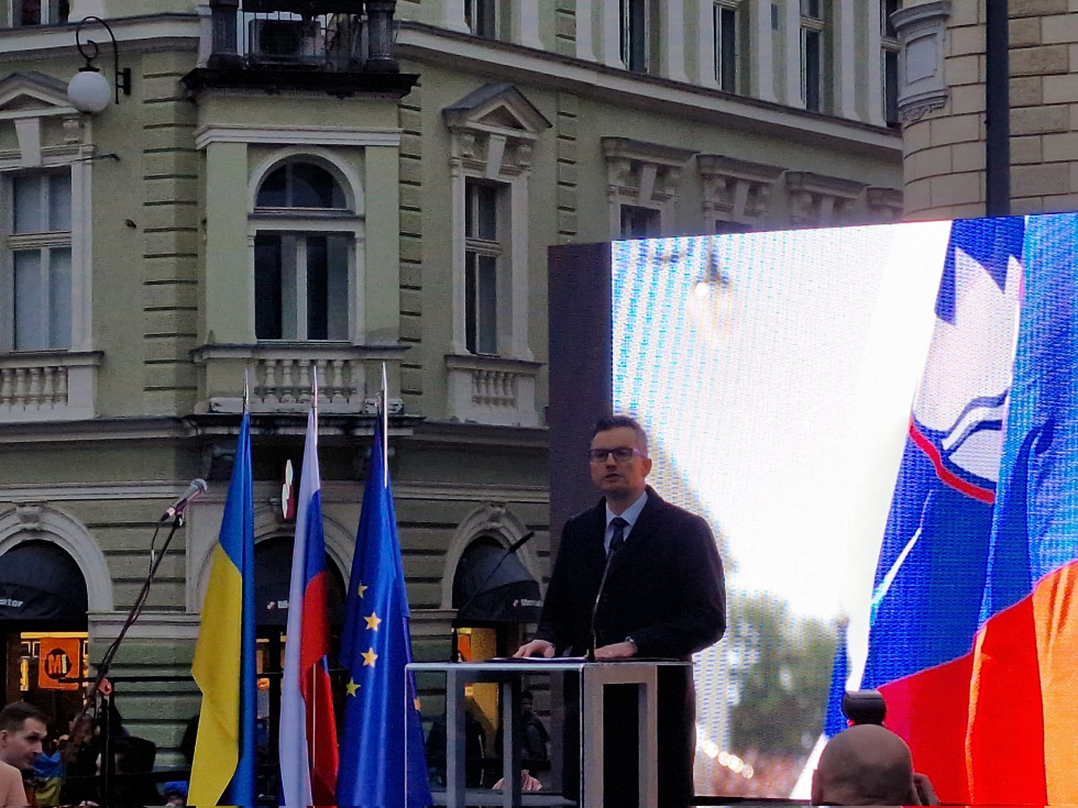 The minister stands on the stage during the speech. Behind him is a large screen and the flags of Slovenia, Ukraine and the EU