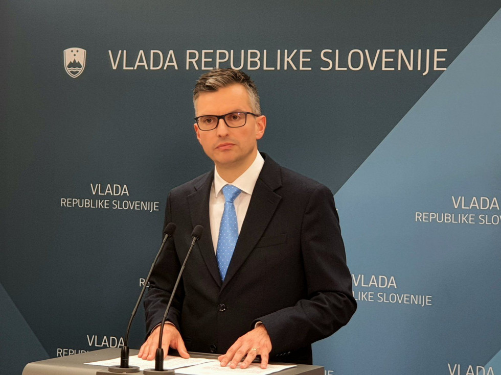 Minister med izjavo stoji za govornico. Za njim je moder pano z logotipom Vlade Republike Slovenije 