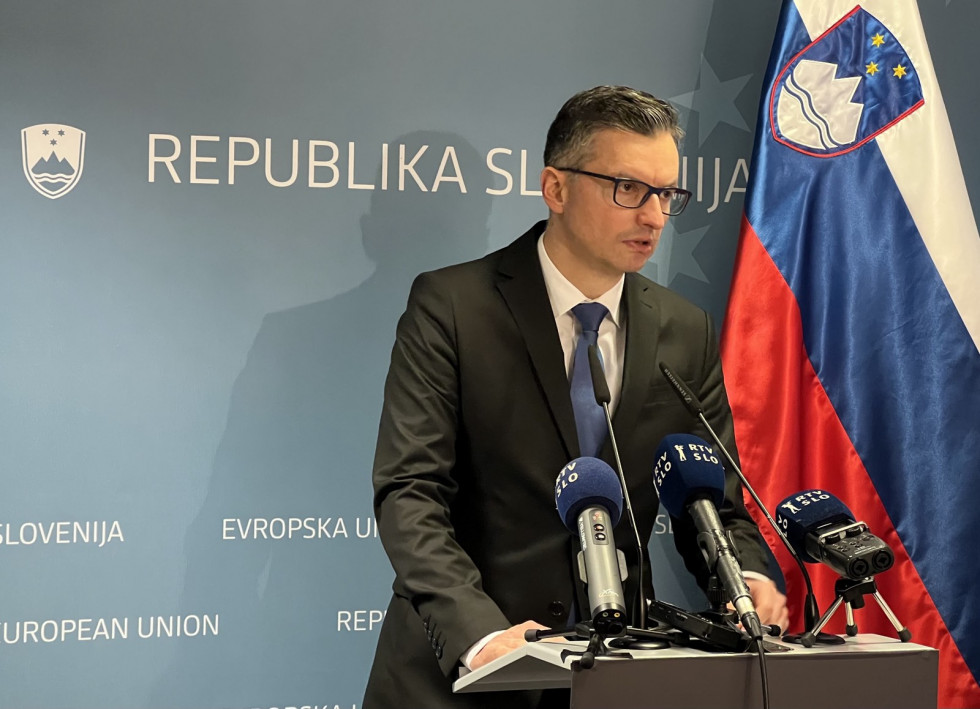 The minister stands behind the lectern during the press conference. Behind is a blue billboard and the Slovenian flag