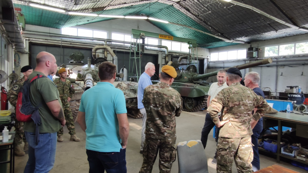 Sekretar in poslanci ter vojaki stojijo v hangarju za oklepna bojna vozila in tanke
