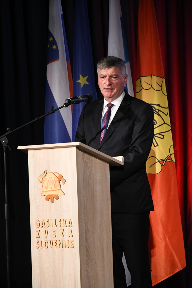 Sekretar med govorom stoji za govornico z znakom gasilske zveze. Za njim sta zastavi gasilske zveze in Slovenije