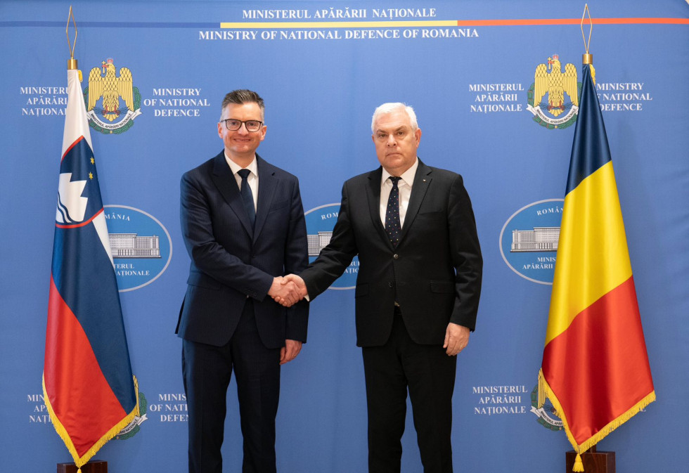Ministra se rokujeta pred modrim panojem. Ob strani sta zastavi Romunije in Slovenije
